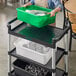 A woman holding a green Vigor bus tub with dishes on top of a cart.