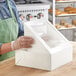 A woman wearing gloves putting a plastic bag of doughnuts into a white Baker's Mark bakery box.