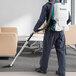 A man wearing a Makita backpack vacuum cleaner vacuuming a room.