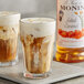 A glass of brown liquid with foam made with Monin Salted Caramel Flavoring Syrup on a table with a bottle of the syrup.