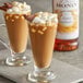 Two glasses of hot chocolate with marshmallows and a bottle of Monin Toasted Marshmallow Syrup on a table.
