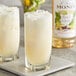 Two glasses of yellow liquid on a tray with a bottle of Monin French Vanilla Syrup.