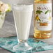 A glass of milkshake next to a bottle of Monin Premium Vanilla Flavoring Syrup.