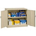 A sand Tennsco storage cabinet with solid doors open to reveal boxes on shelves.