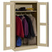 A sand Tennsco wardrobe cabinet with C-Thru doors holding coats and a hat.