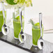 Three Visions silver plastic tasting spoons in glasses of green liquid on a table.