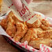 A hand pouring a Mike's Hot Honey packet onto a basket of fried chicken.