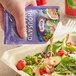 A hand pouring a Kraft Thousand Island dressing packet onto a salad on a table.