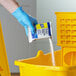 A hand in a blue glove pouring Spic and Span floor cleaner with bleach into a yellow bucket.