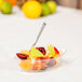 A bowl of fruit salad with a spoon on a clear plastic tray.