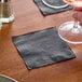 A black 1-ply beverage or cocktail napkin placed on a wooden table.