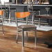 A metal chair with a taupe vinyl seat and a cherry wood backrest, designed for commercial dining settings.
