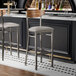 Two Lancaster Table & Seating Boomerang bar stools with a light gray vinyl seat and vintage wood back at a bar counter.