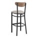 A Lancaster Table & Seating black metal bar stool with a light gray vinyl seat and a vintage wood back.