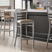 A metal bar stool with a ladder back design and a taupe vinyl padded seat.
