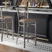 A metal bar stool with a cross back design and a taupe vinyl padded seat.