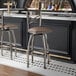 A metal bar stool with a cross back design and a taupe vinyl padded swivel seat.