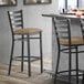 A Lancaster Table & Seating black finish ladder back bar stool with a taupe vinyl padded seat next to a table.