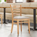 A Lancaster Table & Seating wooden chair with a light gray vinyl cushion next to a table in a restaurant.