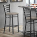 A Lancaster Table & Seating black finish ladder back bar stool with a dark gray vinyl padded seat next to a table.