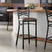 A black metal bar stool with a cross back design and a taupe vinyl padded seat.
