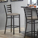 A Lancaster Table & Seating distressed copper ladder back bar stool with a dark gray cushion on the seat next to a table.