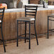A ladder back bar stool with a distressed copper finish and a dark gray vinyl padded seat.