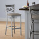 A Lancaster Table & Seating clear coat finish cross back bar stool with a dark gray vinyl padded seat next to a table.