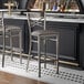 A metal bar stool with a cross back design and a dark gray vinyl padded seat.