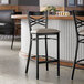 A black metal bar stool with a cross back design and a light gray vinyl padded seat.