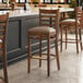 A Lancaster Table & Seating wooden ladder back bar stool with a taupe vinyl seat next to a table with chairs in a restaurant.