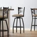 A set of three Lancaster Table & Seating black cross back swivel bar stools with taupe vinyl padded seats.