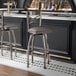 A metal bar stool with a cross back design and a dark gray vinyl padded seat.