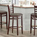 Three Lancaster Table & Seating mahogany wood bar stools with taupe vinyl seats at a wooden bar counter.