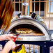 A woman using the WPPO Karma 25 wood fired pizza oven on a patio table to cook a pizza.