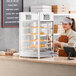 A woman wearing a brown apron and gloves standing behind a ServIt countertop display warmer with food in it.