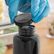 A gloved hand opens a black plastic bottle with a black ribbed cap.
