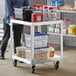 A man in a blue shirt and black pants standing next to a Regency stainless steel utility cart with yellow and red containers on it.