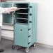 A woman standing next to a blue Cambro double compartment tray carrier in a school kitchen.