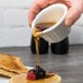 A person pouring brown liquid into a white Tuxton ramekin.