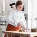A woman in a Uncommon Chef white chef's coat with black piping cutting potatoes.