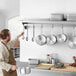A man in a professional kitchen standing in front of a Regency stainless steel wall mounted pot rack with pots and pans hanging from it.