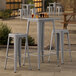 A Lancaster Table & Seating nickel gray outdoor bar height table with two backless barstools on a patio.