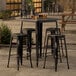 A Lancaster Table & Seating black metal bar table with four stools on an outdoor patio.