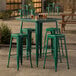 A Lancaster Table & Seating emerald green metal bar table with four backless bar stools on a stone patio.