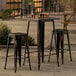 A Lancaster Table & Seating distressed copper bar height outdoor table with 2 backless barstools on a concrete patio.