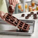 A brown silicone chocolate mold with 15 square compartments being filled with melted chocolate.