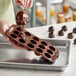A brown silicone chocolate mold with 15 compartments being filled with melted chocolate.