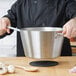 A person holding a Vollrath stainless steel sauce pan over a table with a wooden spoon.