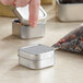 A hand opening a 1 15/16" silver square tin with a slip cover.